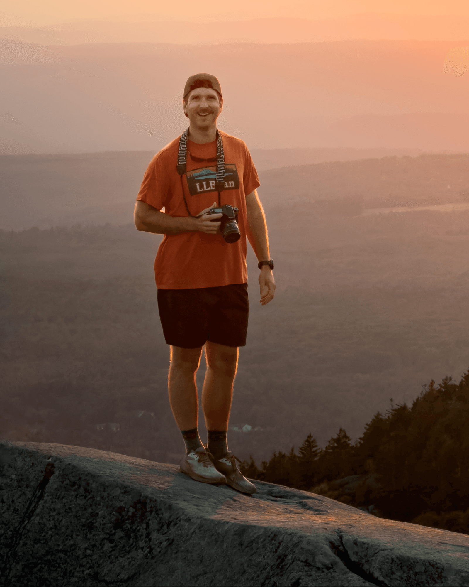 Zach Comeau standing on a rocky overlook with a camera, New England hills behind him